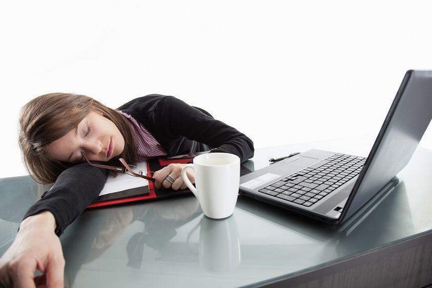 Kobieta śpi na biurku przed komputerem. Ma położoną głowę na jedną rękę pod którą leży notes. Przed nią stoi kawa. A ona śpi...