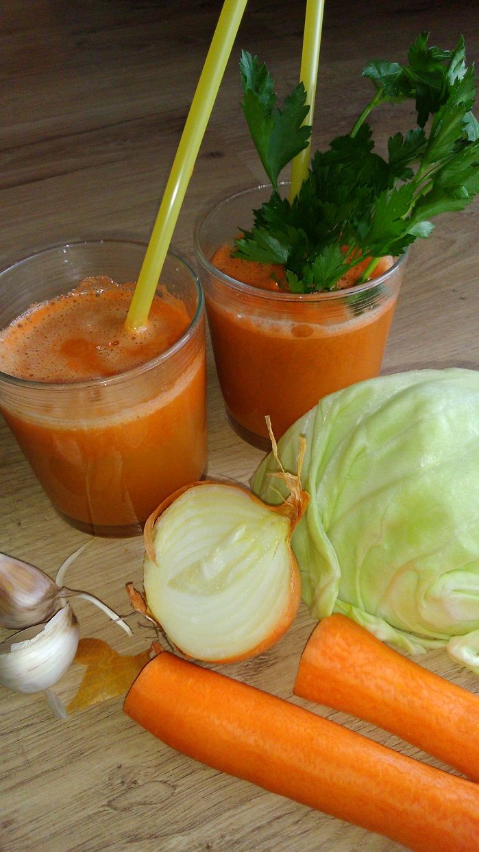 Zdjęcie wyciśniętego soku w dwóch szklankach ze słomką. Obol leżą: dwie marchewki, czosnek, rozkrojona cebulka i kapusta. 