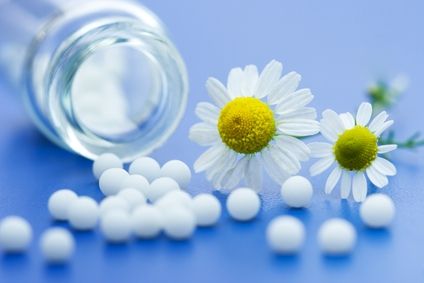 Homeopatczne granulki rozsypane ze szklanej fiolki. Obok dwa kwiatuszki do dekoracji. Na niebieskim tle. 