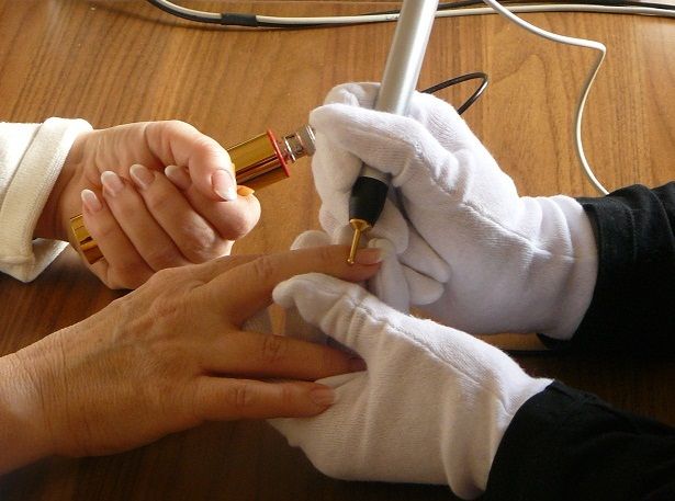 Badanie biorezonansem. Widać dłonie pacjenta i terapeuty. Terapeuta ma rękawiczki i trzyma punktoskop, którym dotyka pacjenta. Dłonie osób są na drewnianym blacie. 